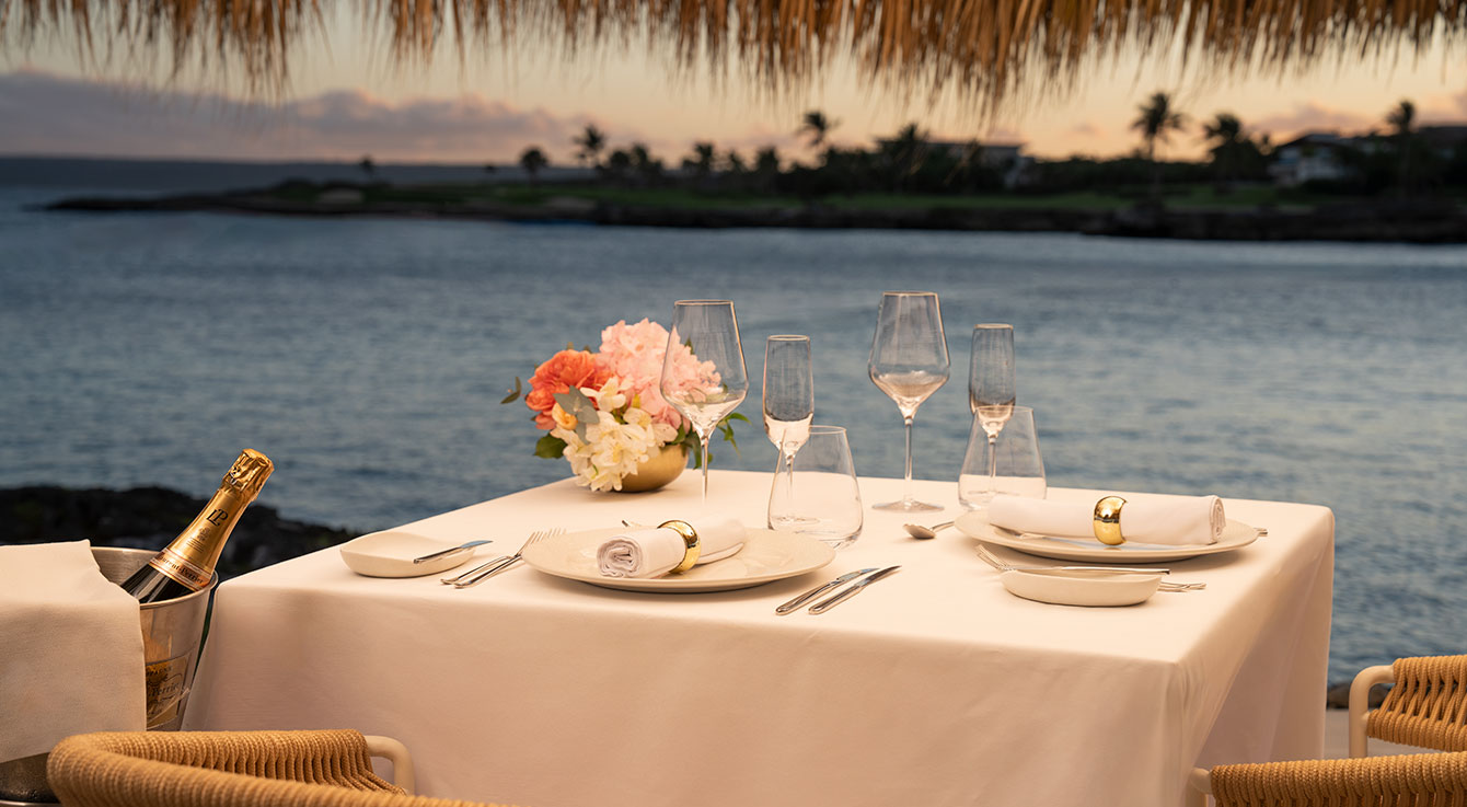 Elegant beachfront dining setup with white tablecloth, champagne, wine glasses, and coral flowers overlooking tropical…