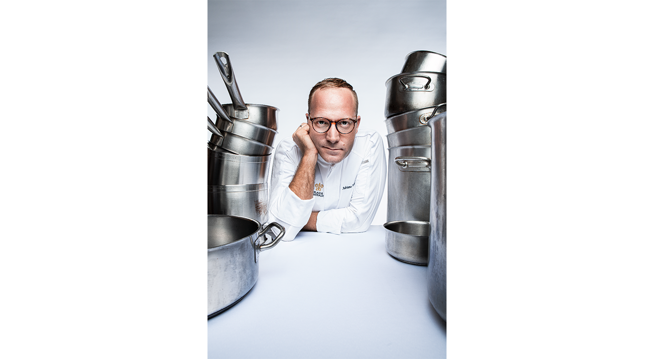 Chef in white coat and glasses seated between stainless steel cooking pots on white surface